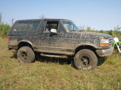 1995 Ford Bronco - A heavily muddied black Ford Bronco sits in a field under a clear blue sky.