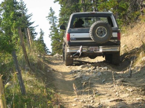 1995 Ford Bronco - A Ford Bronco navigates a challenging off-road trail with trees and a fence line visible.