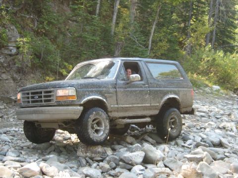 1995 Ford Bronco - A dark gray Ford Bronco navigates a rocky, wooded terrain, with a driver visible behind the wheel.