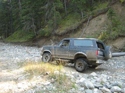 1995 Ford Bronco - Mud-covered 1995 Ford Bronco navigates a rough, rocky path in a forest.