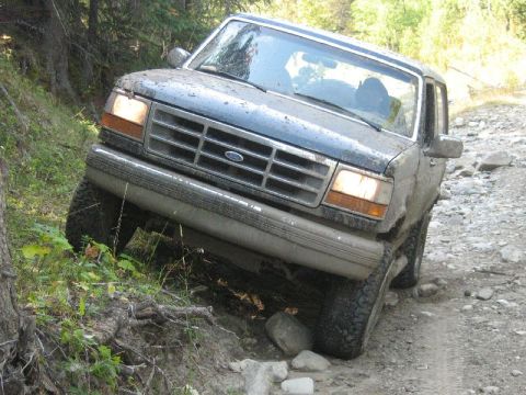 1995 Ford Bronco - A Ford Bronco is shown driving on a rough, unpaved path with trees and rocks surrounding it.