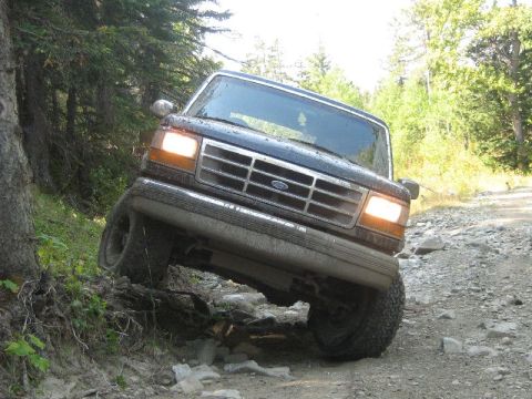 1995 Ford Bronco - A dark blue Ford Bronco navigates a challenging, rocky trail through a forest.