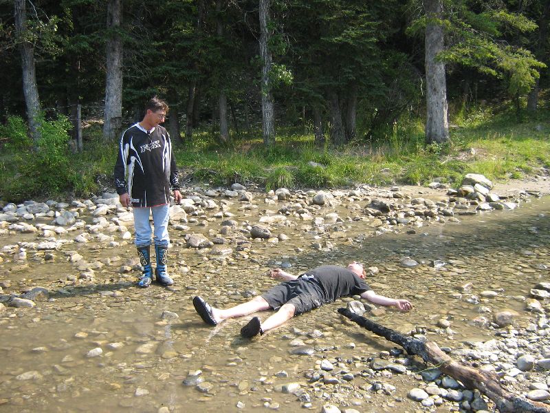 guy in motocross gear watches another guy lie down in a shallow stream bed