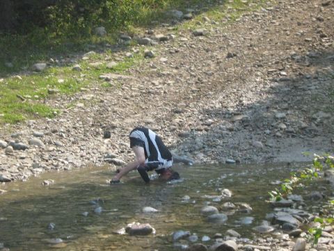 1995 Ford Bronco - A person is bent over in a shallow, rocky stream, with a website logo visible in the lower right corner.