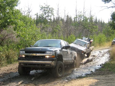 1995 Ford Bronco - A Chevrolet pickup tows a trailer carrying another truck through a challenging muddy path in a wooded area.