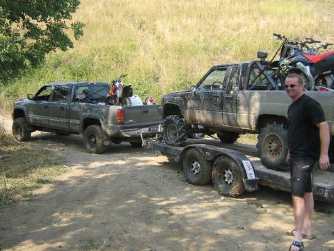 1995 Ford Bronco - A man stands next to a trailer carrying two heavily modified pickup trucks, one of which is loaded onto the other.