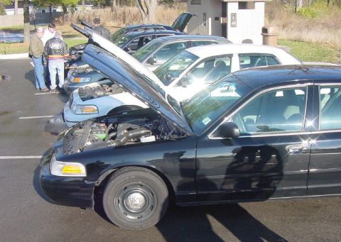 2003 Ford Crown Victoria - Several Ford Crown Victorias, including a black police car with its hood open, are parked in a lot.