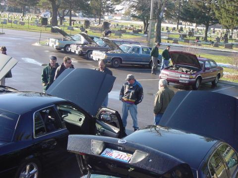 2003 Ford Crown Victoria - A group of car enthusiasts examine vehicles with their hoods up in a parking lot, with a cemetery visible behind them.