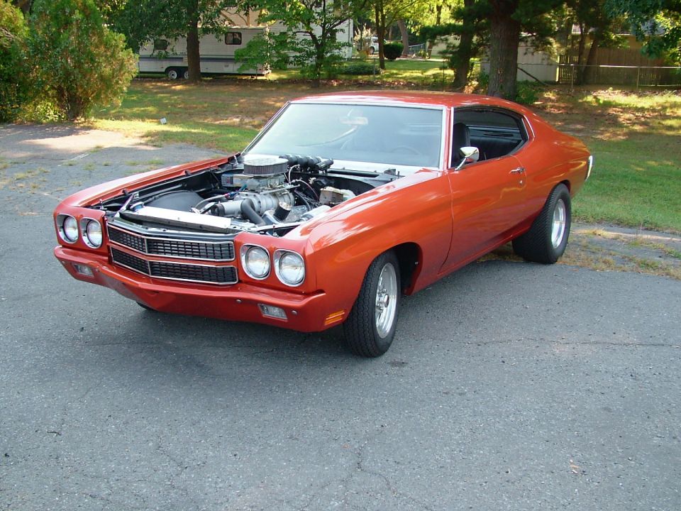 Orange Chevelle coupe with hood off showing blower engine, parked near trees and camper