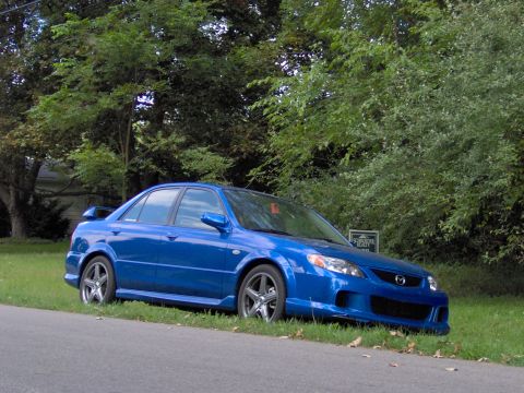 2003 Mazda Protege - A blue 2003 Mazda Protege sedan is parked on the side of the road with trees in the background.