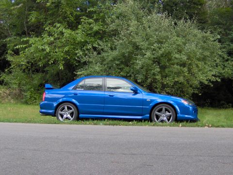 2003 Mazda Protege - This blue 2003 Mazda Protege sedan, equipped with a rear spoiler, is captured in a side profile view parked beside a paved road.