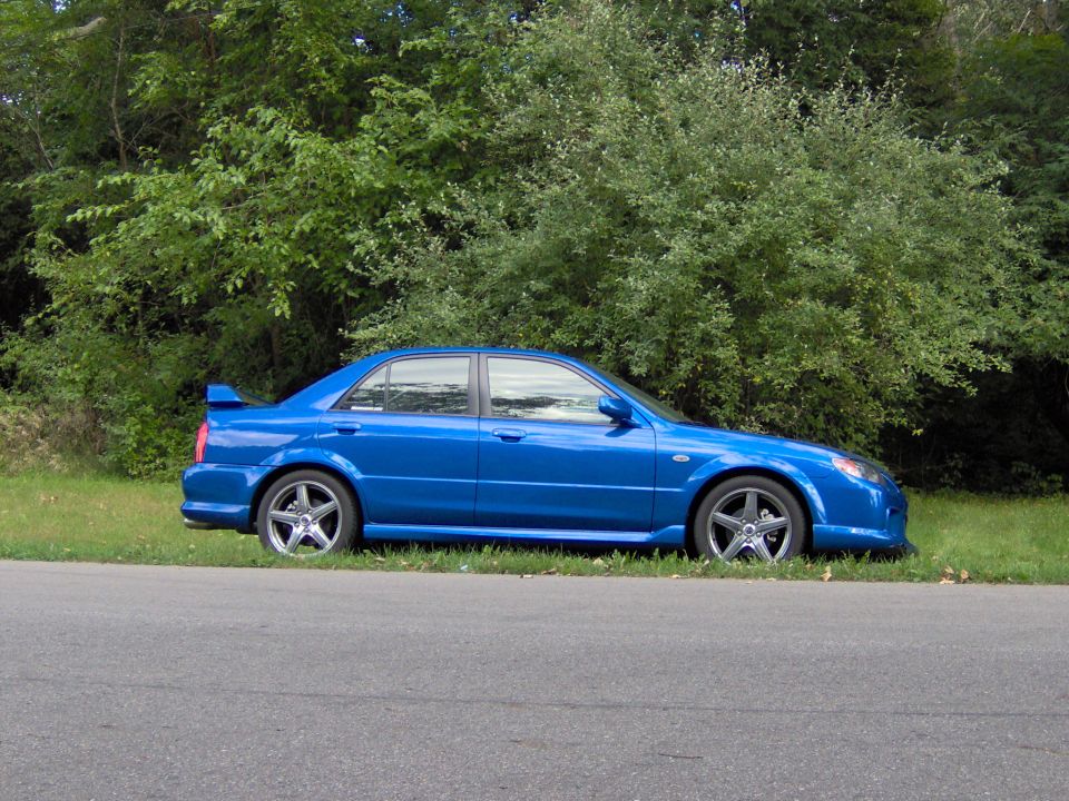 Blue 2003 Mazda Protege sedan with a rear spoiler parked on grass next to a road