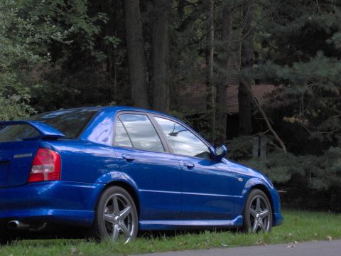 2003 Mazda Protege - A blue 2003 Mazda Protege sedan is parked on a grassy area next to a wooded area.