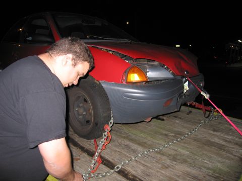 1991 Ford LTD Crown Victoria - A man secures a red car with a damaged front end to a flatbed trailer for transport.