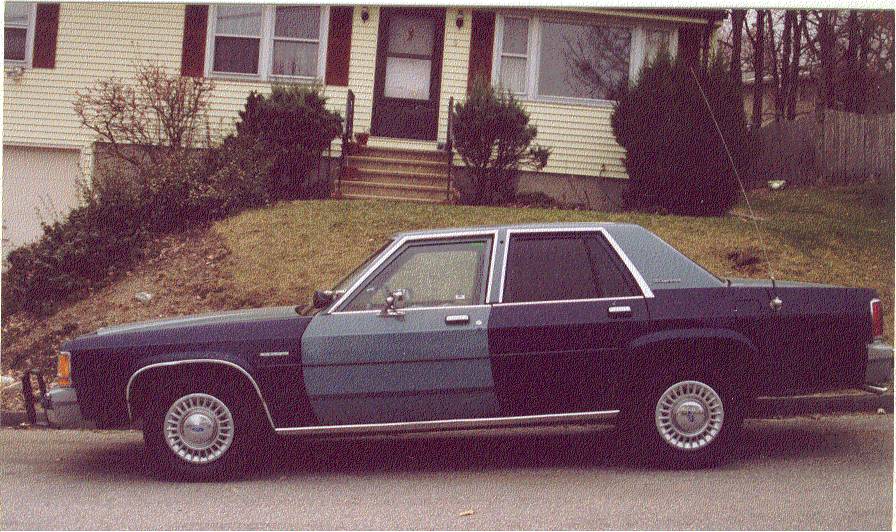 Dark blue 1988 Ford Crown Victoria sedan, side view, parked on street, house in background