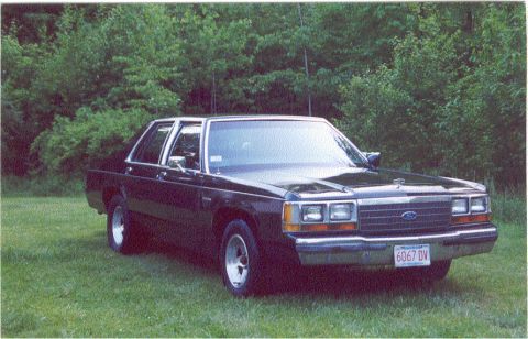 1988 Ford Crown Victoria - A dark-colored sedan is seen from a three-quarter front view, parked outdoors with a grassy area and trees.
