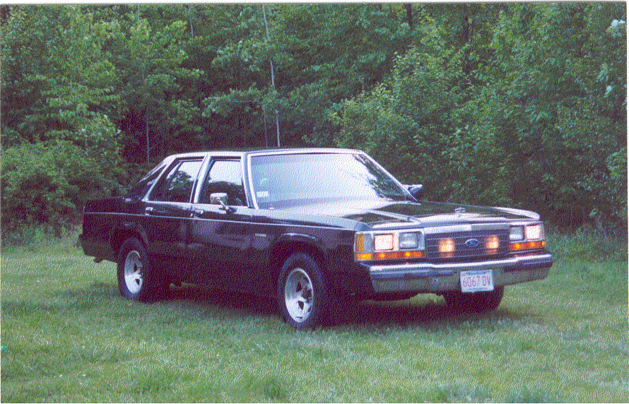 Black boxy Ford Crown Victoria sedan on steel wheels parked on grass before a treeline