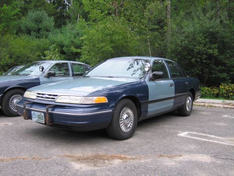 1991 Ford LTD Crown Victoria - A two-tone police car is parked next to another similar vehicle, with trees in the background.