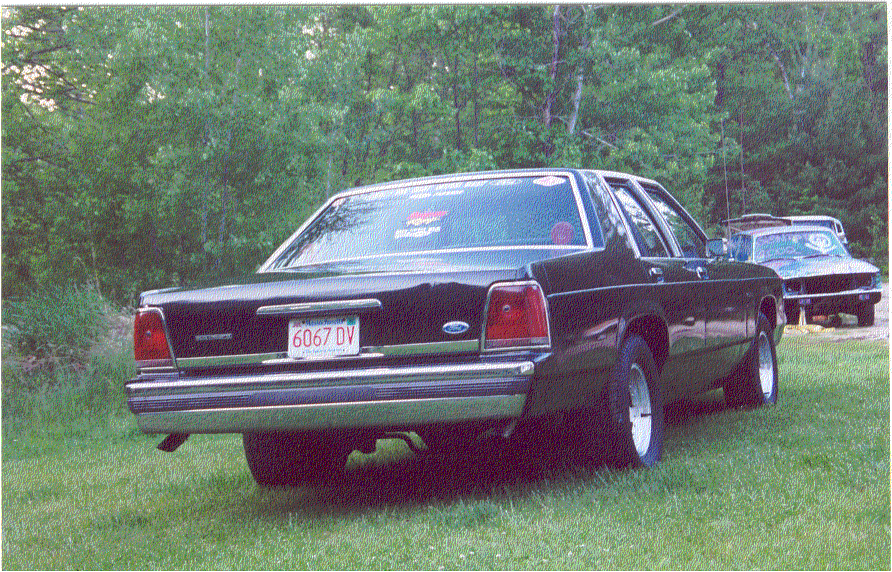 Black boxy Ford sedan on grass, rear three-quarter view with dual exhaust