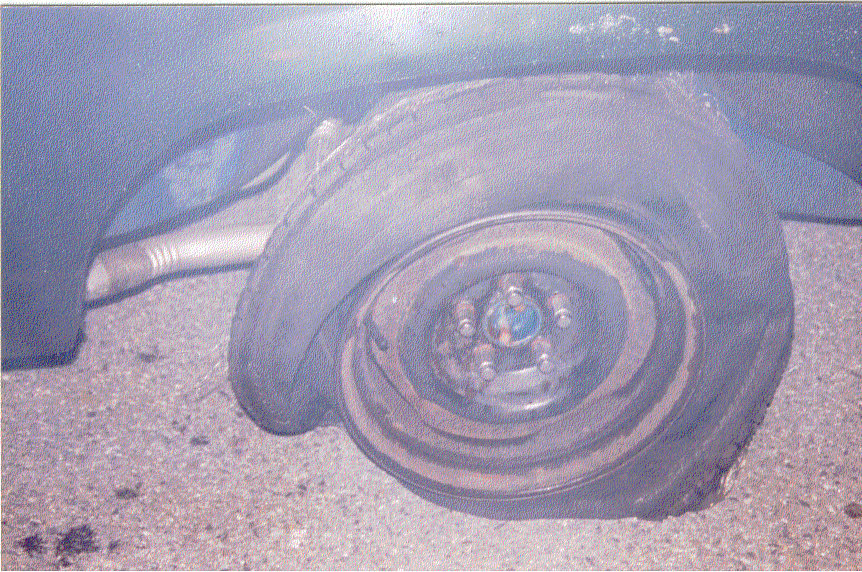 Flat tire on steel wheel with lug nuts under the dark blue fender of a sedan on gravel