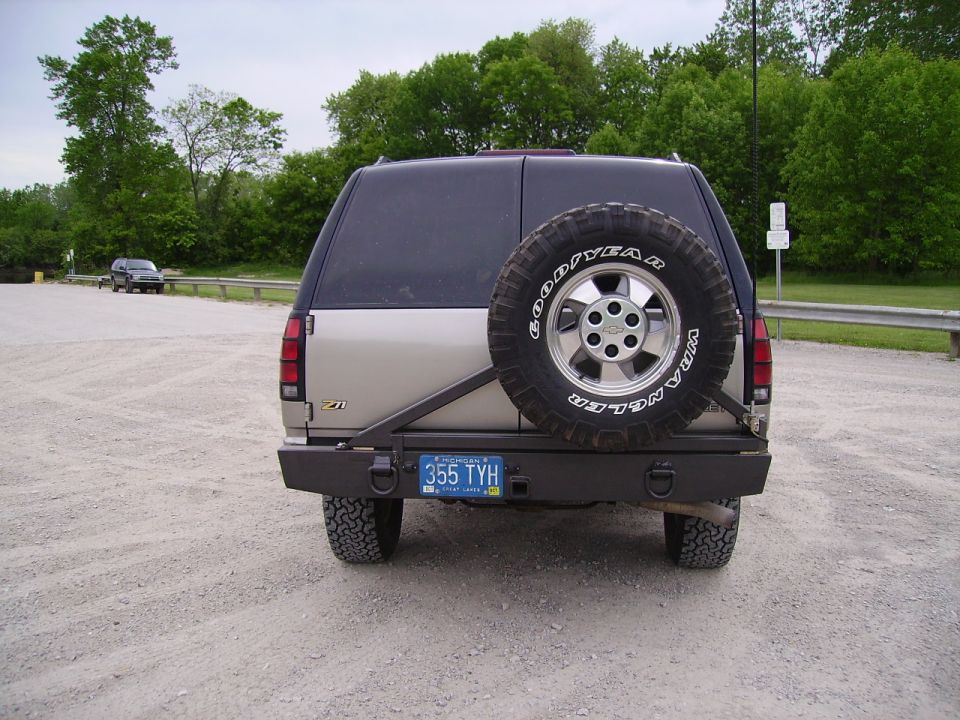 Rear view of a beige and blue SUV with a spare tire mounted on the back. It has a custom bumper and a license plate reading '355 TYH'. Another car is visible in the background