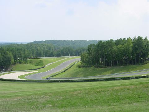 1986 Ford Mustang - A lone dark car navigates a sweeping turn on a race track surrounded by lush green hills and trees.