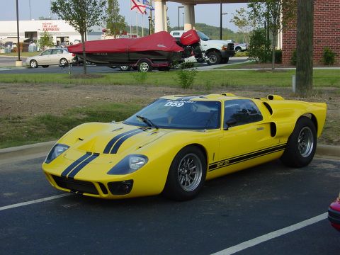 1986 Ford Mustang - A yellow Ford GT40 with black racing stripes is parked in a lot next to a boat on a trailer and a white truck.
