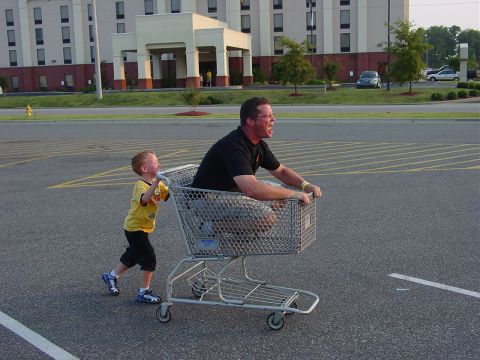 1986 Ford Mustang - A father enjoys a ride in a shopping cart as his son pushes him through a parking lot.
