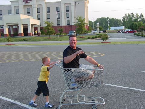 1986 Ford Mustang - A boy pushes a man in a shopping cart in a parking lot outside a Hampton Inn.