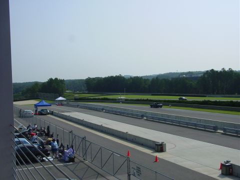 1986 Ford Mustang - Cars gathered at a race track with a vehicle in motion on the course.