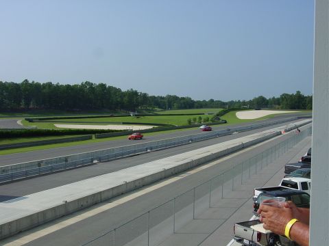 1986 Ford Mustang - Cars race on a sunny day at a track, with a crowd watching from the stands.
