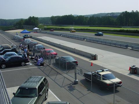 1986 Ford Mustang - A dark coupe with a spoiler is seen on a race track, with several other vehicles parked in the pit area.