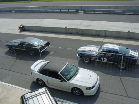 1986 Ford Mustang - Multiple vehicles are visible on a paved track, with a fence in the foreground.
