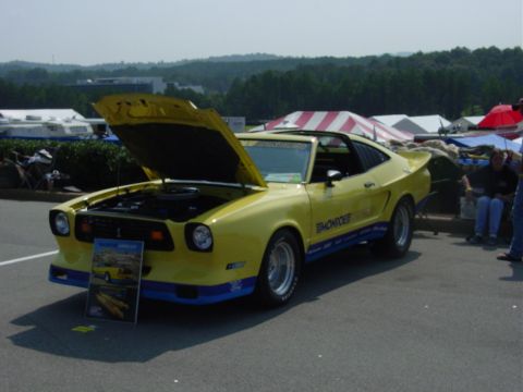 1986 Ford Mustang - A yellow 1977 Ford Mustang II coupe is displayed with its hood open at an outdoor event.