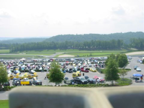 1986 Ford Mustang - A large gathering of vehicles fills a parking lot adjacent to a racetrack, with trees and hills in the background.