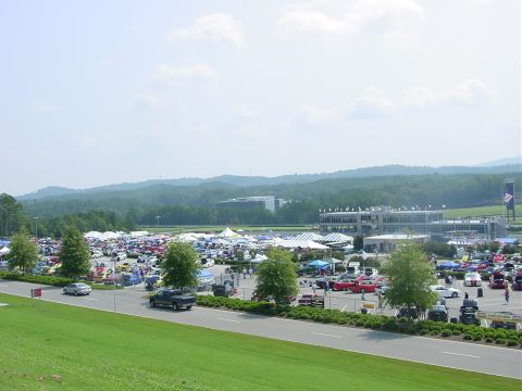 1986 Ford Mustang - A large outdoor gathering features numerous vehicles parked amidst tents and buildings, set against a backdrop of rolling hills.