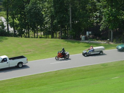 1986 Ford Mustang - A white pickup truck, an orange motorcycle, and a golf cart are traveling down a paved road bordered by grass and trees.