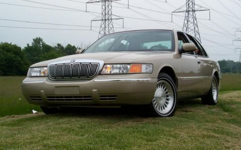 1999 Mercury Marquis - A tan sedan is viewed from the front three-quarter angle, with power lines overhead and grass below.