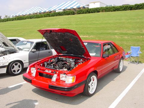 1986 Ford Mustang - A red 1986 Ford Mustang coupe is displayed with its hood open, revealing the engine, alongside another Mustang at an outdoor event.