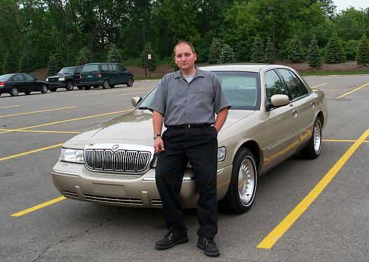 Man leaning on gold Mercury Grand Marquis sedan in empty parking lot