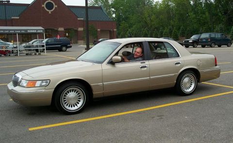 1999 Mercury Marquis - A tan 1999 Mercury Marquis sedan is parked in a lot with a driver visible through the window.