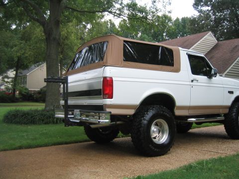 1996 Ford Bronco - White two-tone Ford Bronco with tan lower body and tan soft top, shown from the rear three-quarter angle, lifted on large BFGoodrich mud-te…