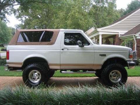 1996 Ford Bronco - A white two-door Ford Bronco with tan lower body cladding and a tan soft top, shown in profile facing right. It sits on a lift kit with ove…