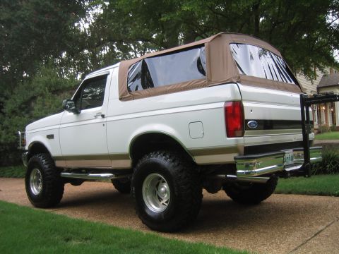 1996 Ford Bronco - White two-tone Ford Bronco with tan/gold lower body accent and a tan soft top, shown from the rear three-quarter angle on a paved driveway…