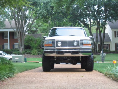 1996 Ford Bronco - Low head-on shot of a lifted white 1990s full-size Ford Bronco sitting on a residential driveway, showing the chrome grille and brush guard…