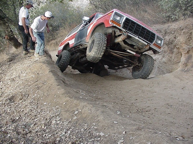 Red-and-white vintage Ford Bronco doing a three-wheel stand in a deep dirt gully as two spotters watch
