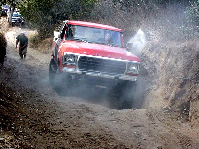 Red 1979 Ford Bronco driving on a dirt trail, kicking up dust. Another vehicle is visible further up the trail