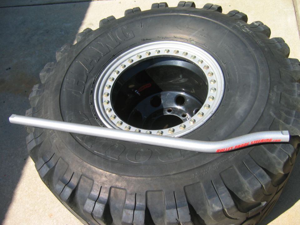 Off-road tire on silver beadlock wheel with a Bullet Proof Steering tie rod laid across it