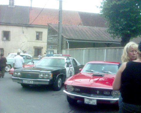 1991 Ford Crown Victoria - A police car and a red muscle car are parked on a street with buildings in the background.