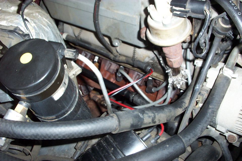 Engine bay close-up with exhaust manifold, gray plug wires, black canister, vacuum actuator and hoses